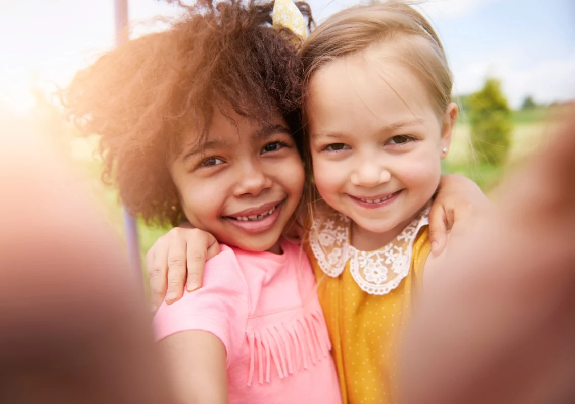 dve devojčice, jedna crne puti druga bele, zagrljene gledaju u fotoaparat za decu i prave fotografiju